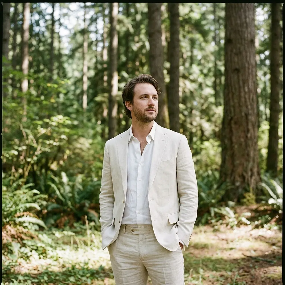 A groom in a linen suit standing in a forest clearing.