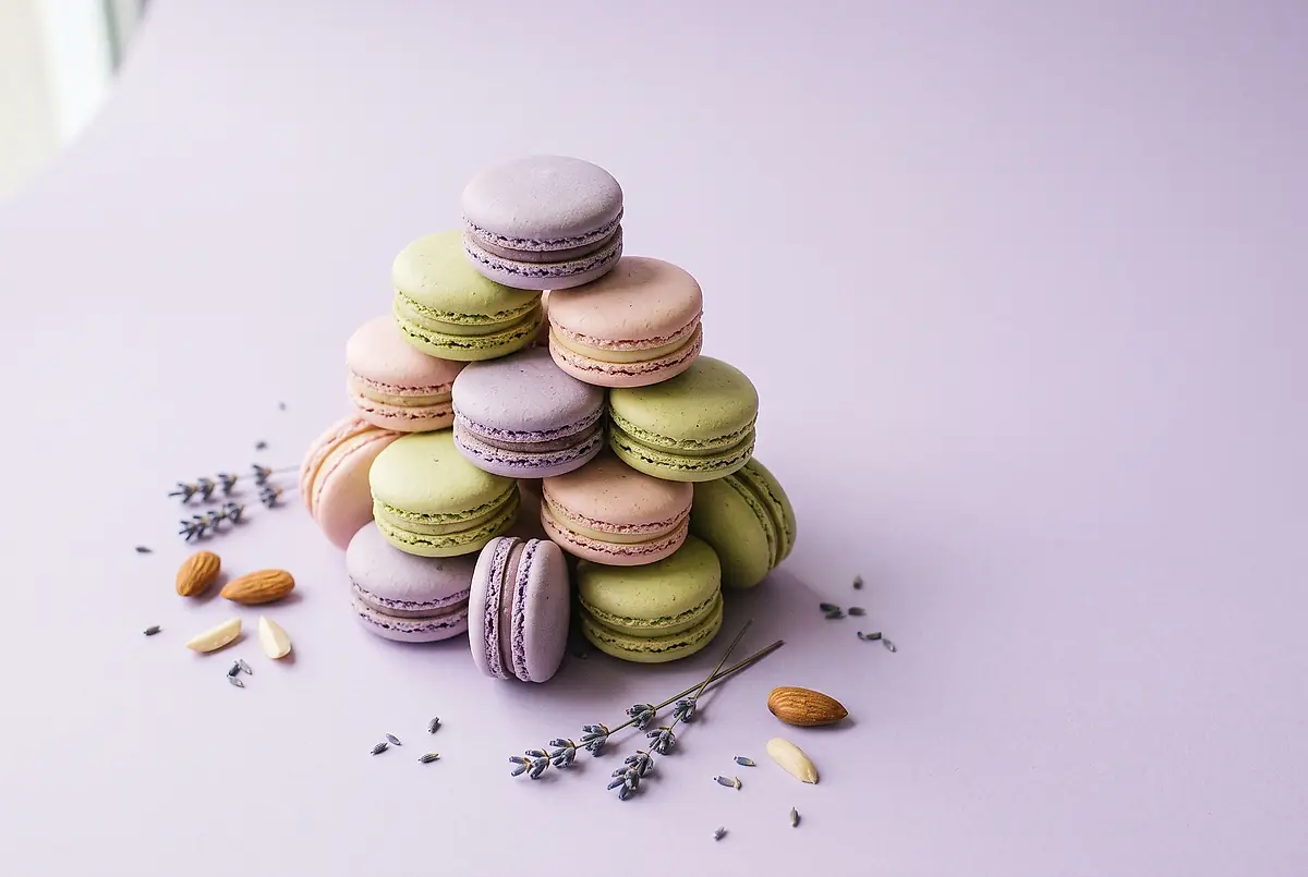 A stack of pastel macarons with scattered almonds and sprigs of dried lavender on a pale lavender table