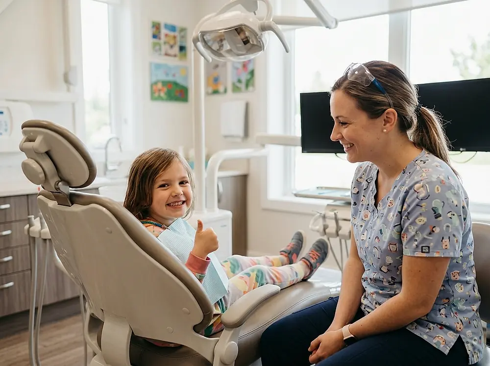 A child giving a thumbs up in a dental chair beside a dental nurse.