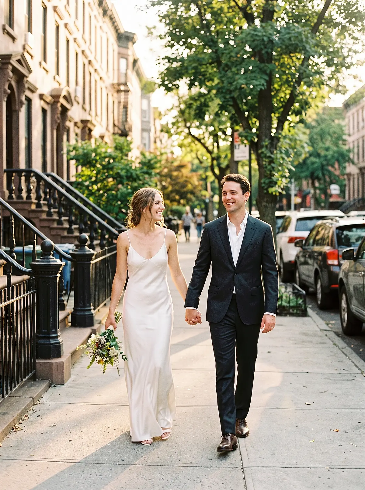 A bride and groom walking hand in hand down a sunlit Brooklyn street of brownstone stoops.
