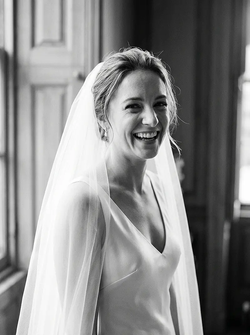 A bride laughing beside a window, photographed in black and white.