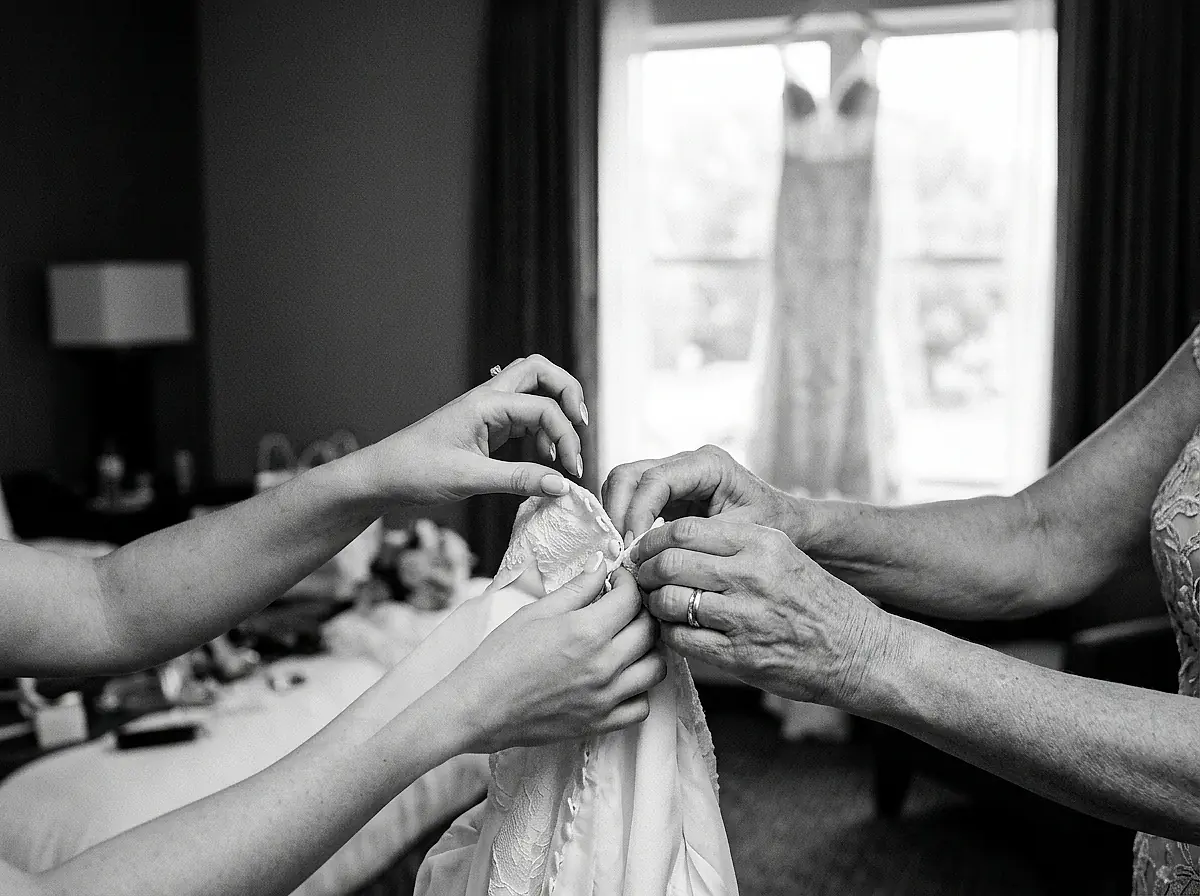 A bridesmaid fastening the back of a bride's dress, photographed in black and white.