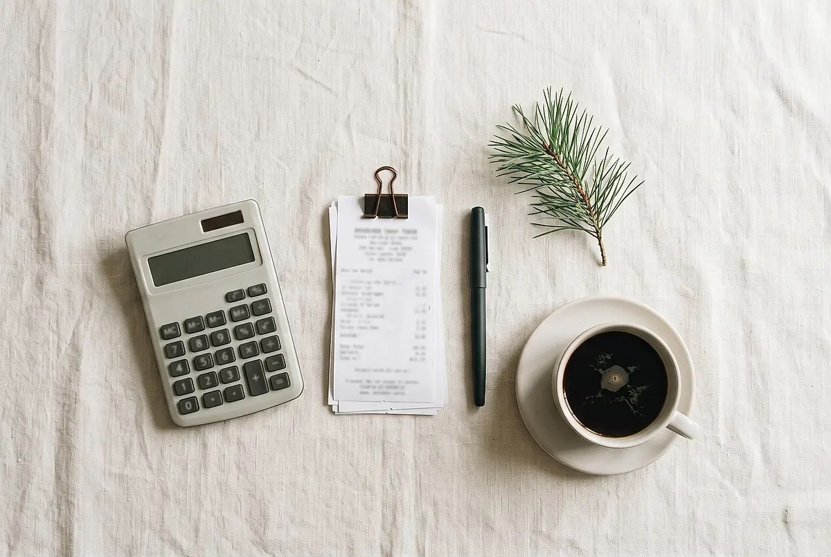 A calculator, a stack of receipts, a pen and a cup of coffee on a pale cloth.