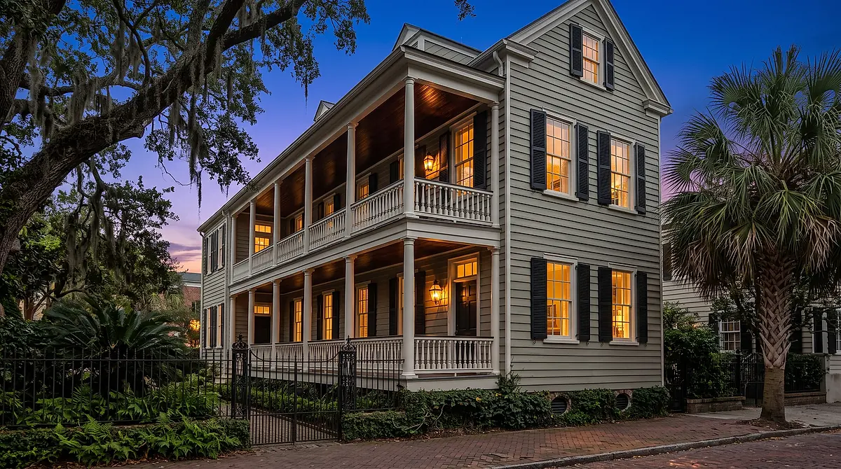 A historic Charleston single house with a two-tier piazza, its windows lit at dusk, behind a live oak in the front garden.