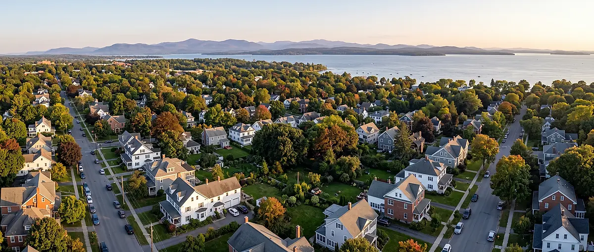 Aerial view of a Burlington neighbourhood: white and grey houses on tree-lined streets, with Lake Champlain and the mountains in the distance