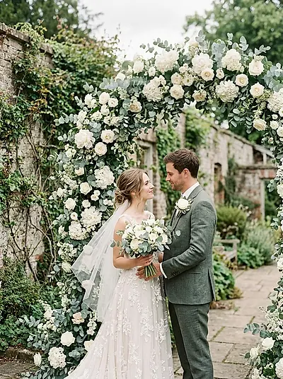 A couple standing beneath an arch of white blooms in a walled garden