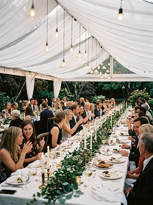 Guests at a long dinner table under a draped white tent