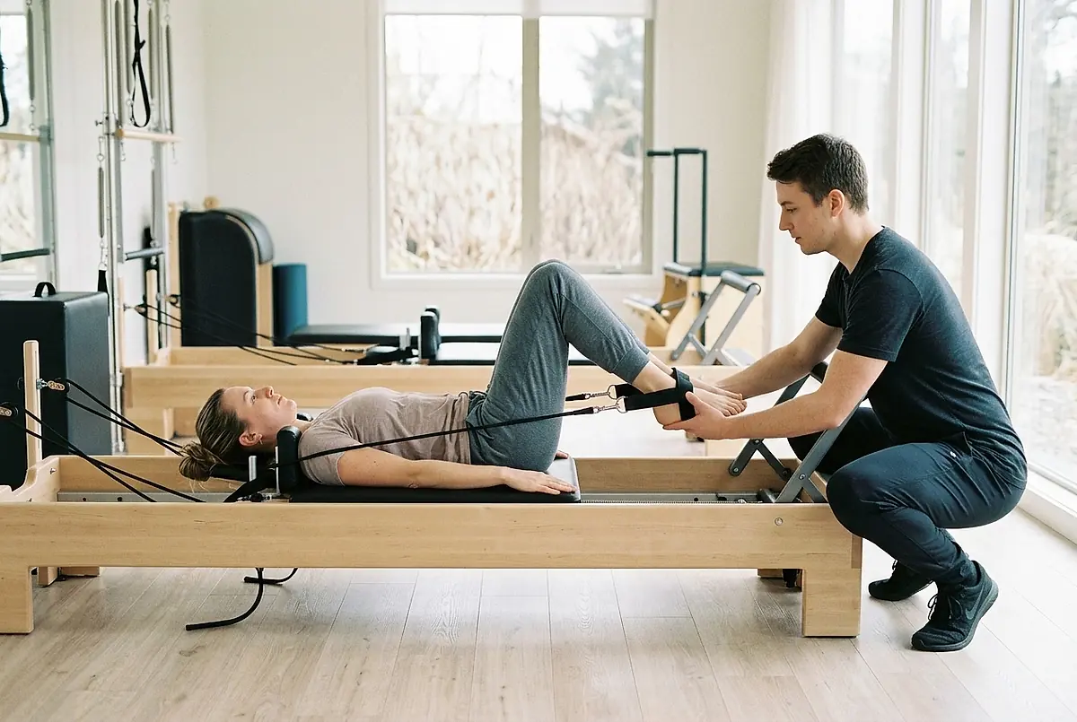 A woman on a Pilates reformer with her feet in the straps while a rehab aide guides her foot.