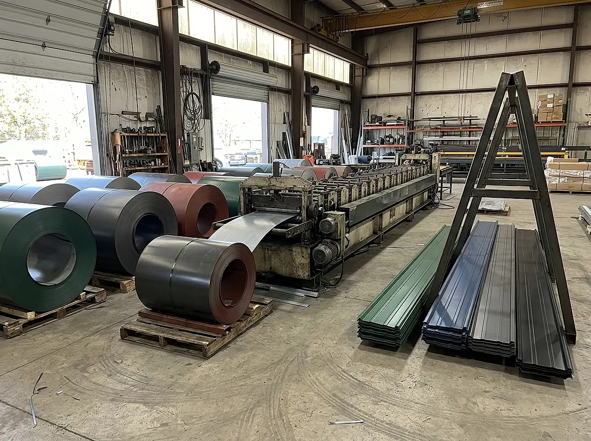Coils of painted steel beside a panel roll forming machine and finished panels in several colors.