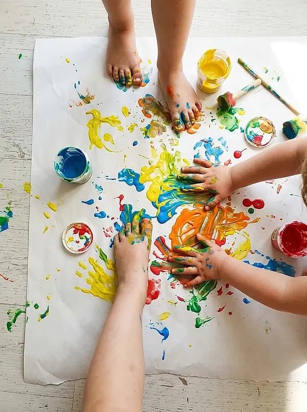 A child's paint-covered hands and feet on a sheet of paper on the floor.