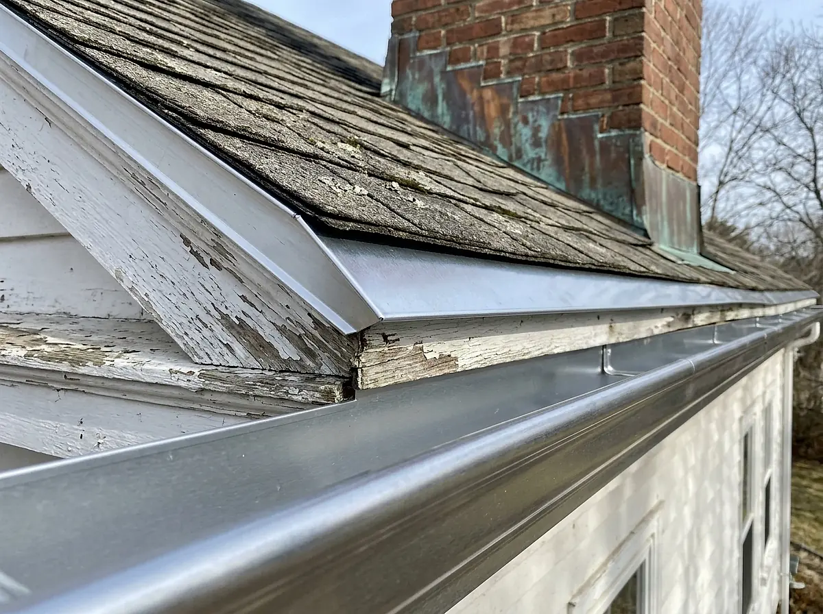 A gutter and drip edge along the eave of an old house, with weathered flashing at the chimney.