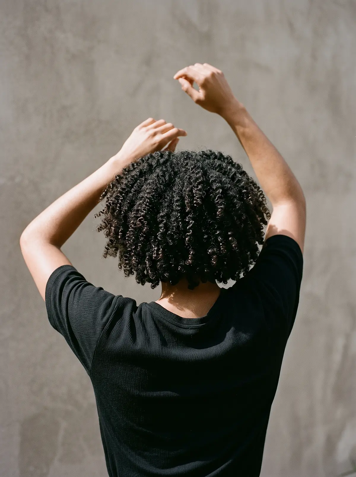 A woman with springy natural coils, seen from behind with her arms raised